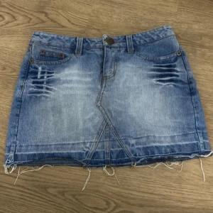 Blå jeanskjol - Snygg blå jeanskjol med slitna detaljer och fransig nederkant. Kjolen har en klassisk femficksdesign och en knappstängning fram. Perfekt för en avslappnad look. Storlek 34 och den är 32cm lång, 47cm nertill(rakt över) och 36cm i midjan(rakt över).