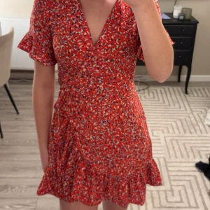 Röd blommig klänning från Retro & Icon - Säljer en söt röd klänning från Retro & Icon med blommigt mönster i blått och vitt. Klänningen har en v-ringad design och korta ärmar med volangdetaljer. Den är även omlott så du kan knyta.  Perfekt för en somrig look! 🌸mycket bra skick storlek xs