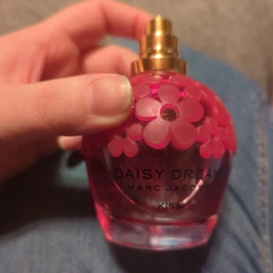 Daisy Dream Kiss parfym från Marc Jacobs - En vacker parfymflaska från Marc Jacobs, Daisy Dream Kiss. Flaskan är klotformad och pryds av rosa blommor som ger en lekfull och feminin känsla. Den har en gyllene spraypump som ger en lyxig touch. Perfekt för den som älskar blommiga och fruktiga dofter. Själva toppen saknas.