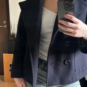 Mörkblå kappa - Säljer en stilren mörkblå kappa med knappar framtill. Jackan har en klassisk design med bred krage och långa ärmar. Perfekt för kyligare dagar och ger en elegant touch till din outfit. Lite nopprig som visas på bilderna annars fint skick.