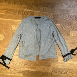 Grå kofta med svarta rosetter - Snygg grå kofta från VERO MODA med långa ärmar och svarta rosetter vid ärmsluten. Perfekt för en stilren look med en twist. Koftan är i en mjuk och bekväm bomullsblandning.