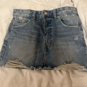 Blå jeanskjol från Pull&Bear - Snygg blå jeanskjol från Pull&Bear i en kort modell. Kjolen har en sliten look med fransiga kanter och klassiska femficksdetaljer. 