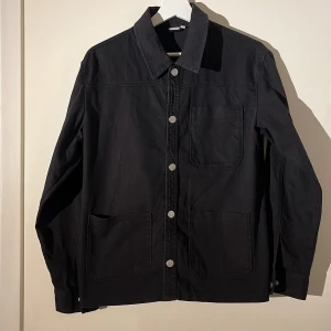 Svart overshirt f - Snygg svart overshirt från SRNHLT med knappar framtill. Den har en klassisk krage och flera praktiska fickor. Perfekt för lager-på-lager-stil och passar till många olika outfits.