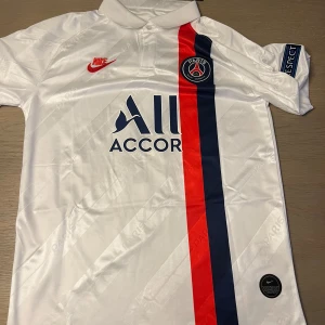 Vit PSG fotbollströja från Nike - Säljer en vit PSG fotbollströja från Nike med korta ärmar. Tröjan har en röd och blå rand på framsidan och PSG-loggan på bröstet. På baksidan finns texten 'NEYMAR JR' och nummer 10. Perfekt för fotbollsfans!
