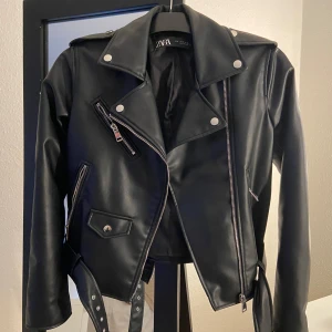 Svart skinnjacka från Zara - Snygg svart skinnjacka från Zara med coola dragkedjedetaljer och knappar. Jackan har bälte i midjan och långa ärmar. Perfekt för att ge din outfit en edgy touch. Den är använd men i väldigt bra skick! 