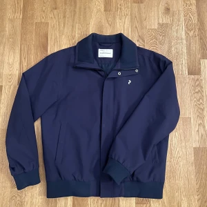 Mörkblå jacka från Peak Performance - Säljer en stilren mörkblå jacka från Peak Performance. Jackan har en dragkedja och knappar framtill, samt ribbade muddar vid ärmslut och krage. Perfekt för en casual look.