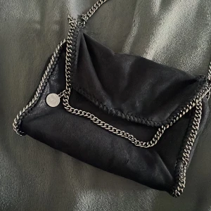 Svart axelväska med kedjedetaljer - Snygg svart axelväska från Stella McCartney med coola kedjedetaljer runt kanterna. Väskan har en stilren design med en rund metallbricka som prydnad. Perfekt för att ge din outfit en edgy touch. Vet inte om den är äkta eller inte men ser ut som det lite smp skadeor här o dör men inget som märks av!