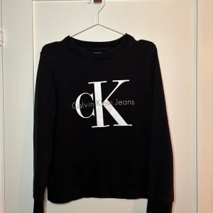 Svart sweatshirt från Calvin Klein | czarna koszula - Snygg svart sweatshirt från Calvin Klein med stort vitt CK-logotryck på framsidan. Tröjan har långa ärmar och en klassisk rund halsringning. 