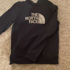 Mörkblå hoodie från The North Face - Säljer en svart hoodie från The North Face med deras ikoniska logga i vitt på bröstet. Tröjan har en klassisk design med långa ärmar och en praktisk magficka.