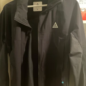 Svart jacka från ACG - Säljer en stilren svart jacka från ACG med dragkedja och hög krage. Jackan har en minimalistisk design med ACG-logga på bröstet. Perfekt för alla väderförhållanden. Knappt använd och därav priset! 10/10 cond. köpt från TRES BIEN i Malmö. 