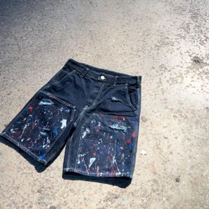 MARES ”PAINTSPLATTER” JORTS - Snygga svarta jeansshorts med unika färgstänk i rött, blått och vitt. Shortsen har en avslappnad passform och slitna detaljer för en cool look. Perfekta för en kreativ och avslappnad stil.