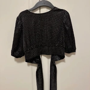 Svart glittrig kort topp med öppen rygg - Snygg svart glittriga topp med prickar och knytband baktill. Toppen har trekvartsärmar och kort i modellen! Aldrig använd. 