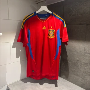 Spanien fotbollströja 2010/2011 Adidas - Säljer en röd kortärmad fotbollströja från Adidas med gula och blå detaljer. Tröjan har ett emblem på bröstet och tre gula ränder på axlarna. Perfekt för fotbollsfans! ⚽