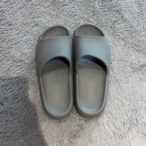 Grå sandaler - Säljer ett par grå sandaler i syntetmaterial. De har en enkel och stilren design med en bred rem över foten. Sulan är mönstrad för bättre grepp. Perfekta för avslappnade dagar.