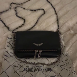 Svart axelväska från Zadig & Voltaire - Snygg svart axelväska från Zadig & Voltaire med kedjerem och coola dragkedjedetaljer. Väskan har en stilren design med en logotyp i form av vingar på framsidan. Perfekt för att ge din outfit en edgy touch. Den är äkta och jag köpte den för 1500kr och säljer för endast 800kr!!😍🙏🏼