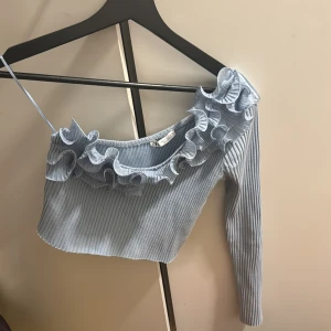 Ljusblå oneshoulder topp från Zara - Säljer en ljusblå oneshoulder topp från Zara med volangdetaljer och ribbad textur. Toppen har en lång ärm och en croppad passform. Perfekt för en trendig look!