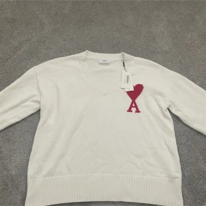 Vit stickad tröja från Ami Paris - Vit stickad tröja från Ami Paris med röd broderad logotyp på bröstet. Tröjan har en v-ringad hals och långa ärmar. Perfekt för en stilren och avslappnad look.