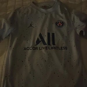 Vit PSG fotbollströja från Jordan - Snygg vit PSG fotbollströja från Jordan med korta ärmar och ett unikt stänkmönster. Tröjan har PSG-loggan på bröstet och texten 'Accor Live Limitless' samt 'Visit Rwanda'. Perfekt för alla PSG-fans! I storlek 158 cm! 