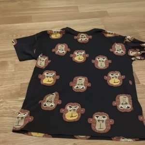 Svart t-shirt med apmönster från Lindex - Cool svart t-shirt från Lindex med ett lekfullt apmönster i brunt och gult. Perfekt för barn som gillar djurmotiv. T-shirten har korta ärmar och en bekväm passform.