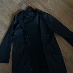 Svart regnjacka från Rains - Säljer en stilren svart parka regnjacka från Rains. Jackan har långa ärmar och en praktisk huva, perfekt för regniga dagar. Den är designad med knappar framtill och har en klassisk passform. 