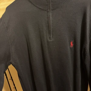 Svart tröja med dragkedja från Ralph Lauren - Säljer en stilren svart tröja från Ralph Lauren med en liten dragkedja vid halsen. Tröjan har en broderad röd logotyp på bröstet och är perfekt för en avslappnad look. Passar bra till både jeans och chinos.