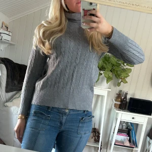 Grå stickad polotröja - Säljer en mysig grå stickad polotröja med kabelstickat mönster. Tröjan har långa ärmar och en hög polokrage som håller dig varm under kyliga dagar. Perfekt för höst och vinter! Super len och mysig, inte alls sticksig