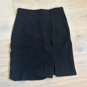 Svart kjol med slits - Snygg svart kjol med en diskret slits framtill. Kjolen har en enkel och stilren design med en elastisk midja för extra komfort. Perfekt för en chic och minimalistisk look. Lappen är bortklippt men skulle gissa på storlek xs/s. En bit an sömmen på nedre delen av kjolen har samt gått upp men är lätt att fixa. Annars har den inga större defekter och är som ny.🥰