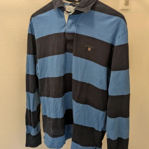 Randig pikétröja från GANT - Säljer en snygg randig pikétröja från GANT i blått och svart. Tröjan har långa ärmar och en klassisk krage. Perfekt för en avslappnad stil med en touch av elegans. Passar bra till jeans eller chinos.