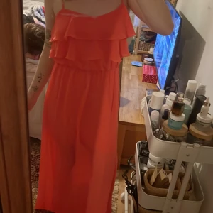 Korallfärgad klänning med volanger - Säljer en snygg orange långklänning med volanger upptill. Klänningen har smala axelband och en slits på sidan som ger en elegant touch. Perfekt för sommarens festligheter! Står st M men jag tycker den sitter thight på mig som har S