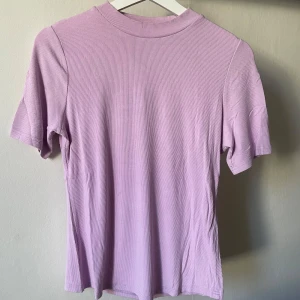 Ljuslila ribbad topp - Säljer en ljuslila ribbad topp med korta ärmar. Perfekt för en avslappnad stil. Toppen har en enkel design med rund halsringning.