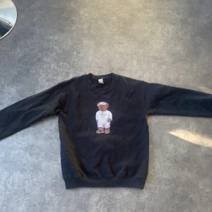 Svart sweatshirt med nalletryck från Gildan - Säljer en svart sweatshirt från Gildan med ett gulligt nalletryck på framsidan. Tröjan är tillverkad i en bomulls- och polyesterblandning och har långa ärmar. Perfekt för en avslappnad stil! 🐻