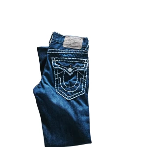 True Religion jeans  - Snygga blå jeans från True Religion med bootcut-stil. De har en unik broderad design på bakfickorna och en knappdetalj. Perfekta för en avslappnad look med en touch av stil.