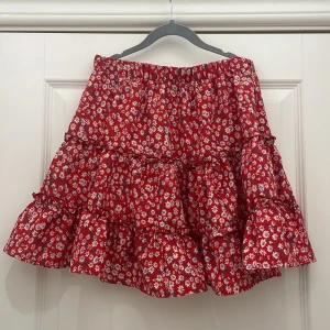 Röd blommig volangkjol - Charmig röd kortkjol med blommigt mönster och volanger. Perfekt för en somrig look med sin livfulla färg och lekfulla design. Kjolen har en elastisk midja för bekväm passform.