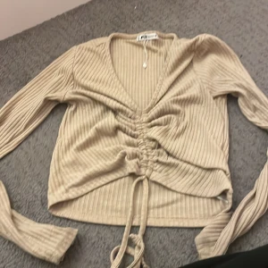 Beige ribbad tröja med knyt - Säljer en beige ribbad tröja med v-ringning och knyt framtill. Tröjan har långa ärmar och är perfekt för en stilren look. Passar bra till både jeans och kjol.