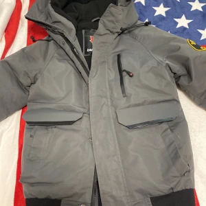 Grå jacka från Zavetti Canada - Säljer en grå jacka från Zavetti Canada med broderad logga på ärmen. Jackan har dragkedja och flera fickor med lock. Perfekt för kyliga dagar med sin robusta design.