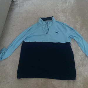 Blå och svart tröja från Puma - Säljer en stilren blå och svart tröja från Puma med dragkedja vid halsen. Tröjan har långa ärmar och en broderad Puma-logga på bröstet. Perfekt för en sportig look!