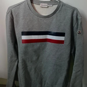 Grå sweatshirt från Moncler - Säljer en stilren grå sweatshirt från Moncler med ett färgglatt band i blått, vitt och rött på bröstet. Tröjan har långa ärmar och rund halsringning. Köptes i New York och blev för liten snabbt, så väldigt oanvänd!