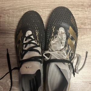 Adidas fotbollsskor med guldiga detaljer - Säljer ett par Adidas fotbollsskor med svart och guldigt mönster. Skorna har en unik textur på ovansidan och snörning för bra passform. Perfekta för fotbollsplanen!