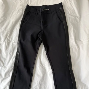 Svarta byxor med läderimitation i sidorna - Snygga svarta byxor från House of Lola med läderimitation längs sidorna. Endast använda 2 gånger. Byxorna har en knapp och dragkedja framtill. Dekorativa bakfickor. Tillverkade i en blandning av bomull, polyamid och elastan för en bekväm passform. Byxorna är i croppad längd och med medelhög midja.
