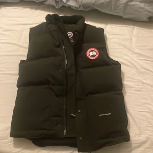 Svart dunväst från Canada Goose - Säljer en svart dunväst från Canada Goose med broderad logga på bröstet. Västen har dragkedja och knappar framtill, perfekt för kyliga dagar. Den är tillverkad i canada goose och har en stilren design. Har aldrig andvänds är som ny 