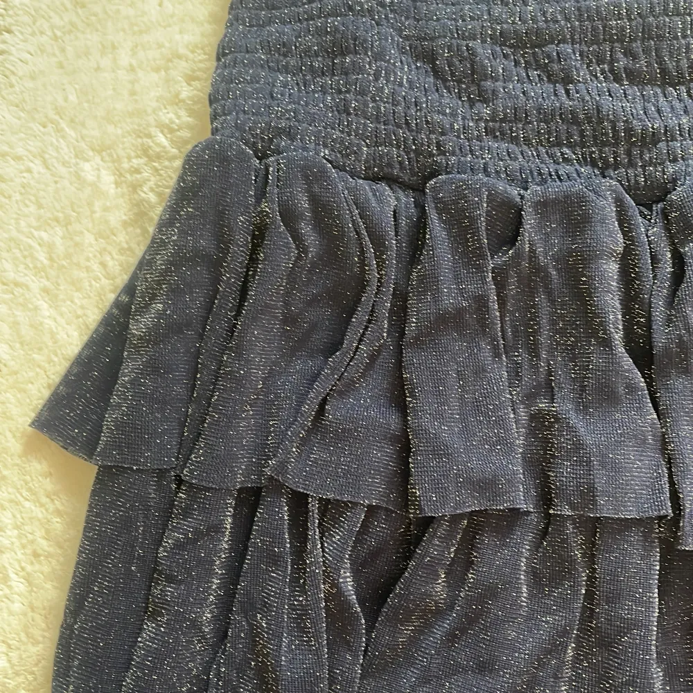 Snygg mörkblå kjol från Neo Noir med glittrande detaljer. Kjolen har en volangdesign som ger en fin rörelse och en elastisk midja för bekväm passform. Perfekt för att lägga till lite glitter i garderoben!. Hameet.