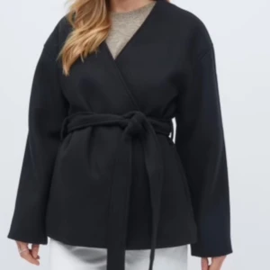 Svart omlottjacka med knyt - Snygg svart omlottjacka med knytband i midjan. Jackan har en stilren design med långa ärmar och en v-ringad hals. Perfekt nu till våren. Jackan är i nyskick, prislapp kvar och nypris 700kr