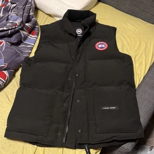 Svart dunväst från Canada Goose - Säljer en svart dunväst från Canada Goose med en broderad logotyp på bröstet. Västen har en dragkedja och knappar framtill samt två stora fickor. Perfekt för kyligare dagar. Västen är köpt för 1 år sedan och har bara legat endast provad. Den ser precis ut som ny. Priset går att diskutera vid snabb affär 