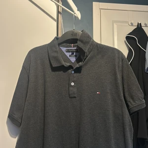 Grå pikétröja från Tommy Hilfiger - Säljer en stilren grå pikétröja från Tommy Hilfiger i slim fit. Tröjan har en klassisk krage och knappar framtill. Perfekt för en avslappnad men ändå snygg look.