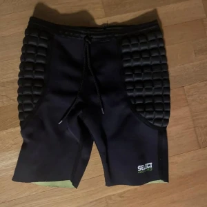 Svarta vadderade målvaktsbyxor från Select - Svarta shorts från Select med vadderade paneler på sidorna för extra skydd. De har en snörning i midjan och är tillverkade av polyamid och neoprenblandning, vilket gör dem perfekta för sportaktiviteter. Storlek M