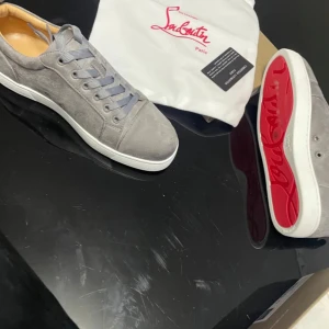 Grå sneakers från Christian Louboutin - Snygga grå sneakers i mocka från Christian Louboutin med den ikoniska röda sulan. Skorna har snörning och en stilren design som passar perfekt för en trendig look.