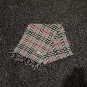 Grå rutig halsduk från Burberry - Snygg grå halsduk från Burberry med klassiskt rutmönster i svart och rött. Tillverkad i mjuk lammull och har fransar i ändarna. Perfekt för att hålla dig varm och stilren under kyliga dagar.