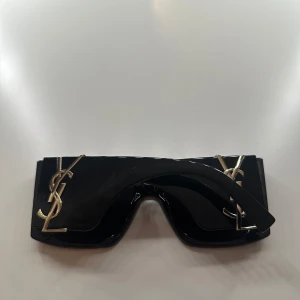 Svarta solglasögon med gulddetaljer - Snygga svarta solglasögon med breda bågar och stiliga gulddetaljer på sidorna. Perfekta för att ge en lyxig touch till din outfit. De har en modern och djärv design som verkligen sticker ut.