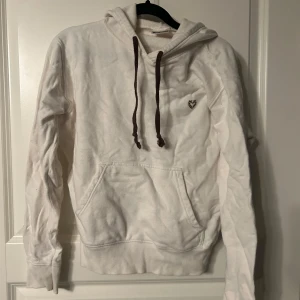 Vit hoodie med bruna snören - Vit hoodie med bruna snören och en liten broderad detalj på bröstet. Den har en klassisk känguruficka framtill och en bekväm huva. Perfekt för en avslappnad stil. Är väldigt liten i storlek skulle säga är som en S