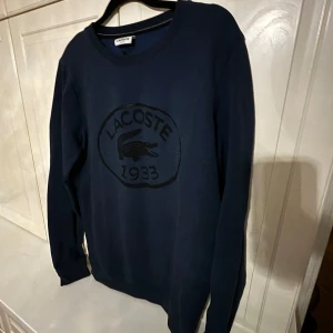 Mörkblå tröja från Lacoste - Säljer en stilren mörkblå tröja från Lacoste med ett stort svart tryck av Lacoste-loggan. Tröjan har en slim fit passform och långa ärmar, Mycket bra skick, storlek S!